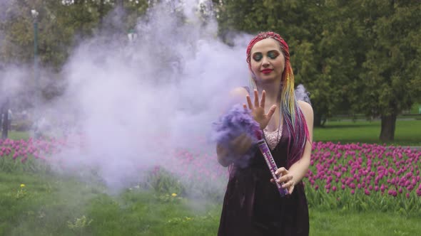 A Girl with a Smile with Bright Makeup and Colored Braids in a Purple Dress Blows Artificial Purple