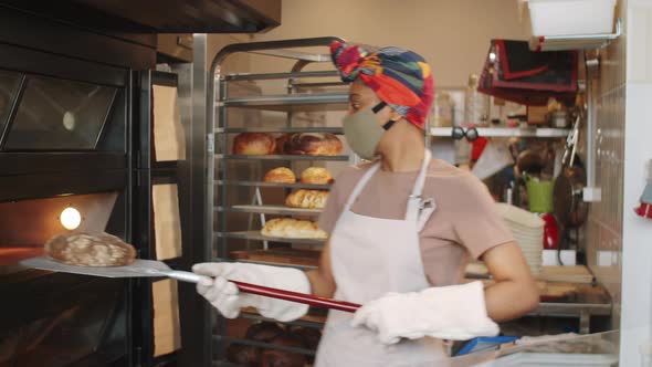 Afro-American Female Baker in Mask Taking Bread from Oven with Shovel