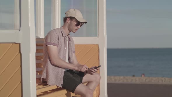 Man Sat On Bench Near Sea Making A Purchase On His Phone - Ungraded