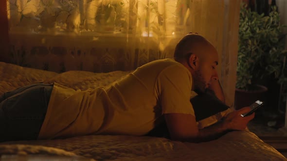 A bald, bearded, bored adult man is lying on a bed and using a smartphone. Rest at home.