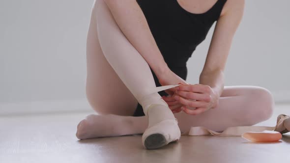 Caucasian female ballet dancer sitting on the floor in the studio and tying her ballet shoes