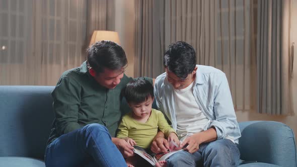 Modern Married Gay Couple Spending Time With Their Son Sitting On Sofa In Living Room Reading Book