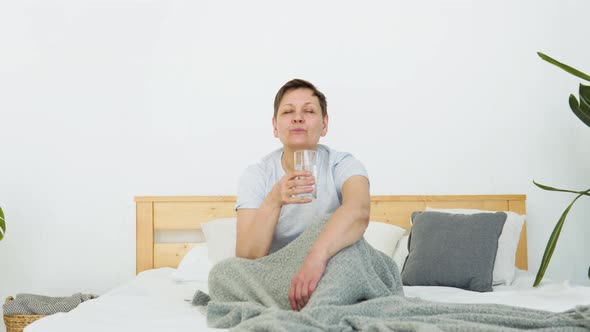 Smiling Beautiful Senior Adult Woman Drinks Water Sitting in Bed