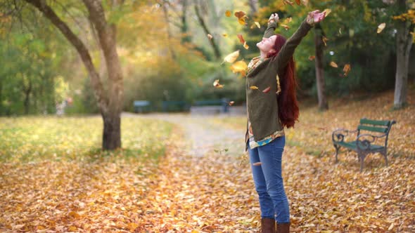 Slow Motion: Beautiful young redhead woman standing in a park in autumn with leaves falling down aro