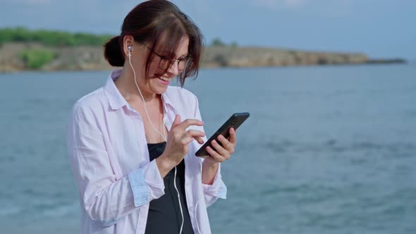 Happy Middle Age Woman Relaxing on Beach Wearing Headphones with Smartphone