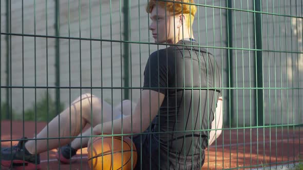 Back Angle View of Relaxed Young Caucasian Basketball Player Sitting on Outdoor Court and Resting