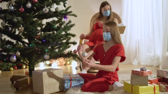 Slim Teenage Girl in Covid Face Mask Decorating New Year Tree with Blurred Mother Reading Book