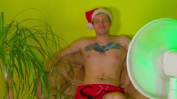 Happy Man in Santa Hat with Bare Torso Sitting in Armchair Before Fan