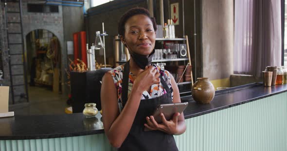 Portrait of african american woman in face mask working at a bar, using tablet and smiling to camera
