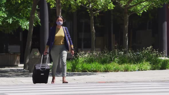 African american woman wearing face mask crossing road wheeling suitcase