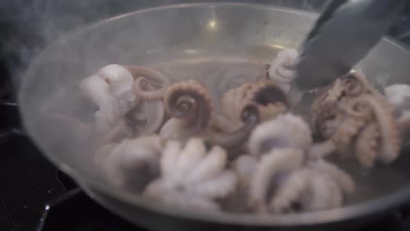 Stirring Octopuses in Metal Frying Pan with Tongues in Kitchen