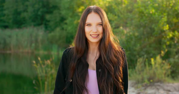 Portrait of a Smiling Young Woman with Long Dark Hair Outdoors in a Forest Near a Lake