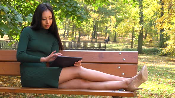 A Young Asian Woman Sits on A Bench with Legs Stretched out In a Park and Works on A Tablet