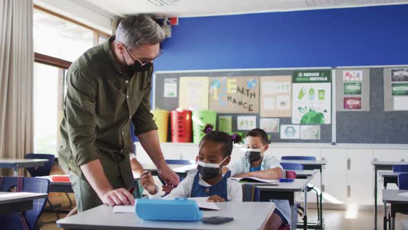 Diverse male teacher helping schoolgirl sitting in classroom, all wearing face masks