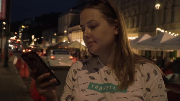 Young Girl Writes a Message on the Phone Against the Background of a City Street