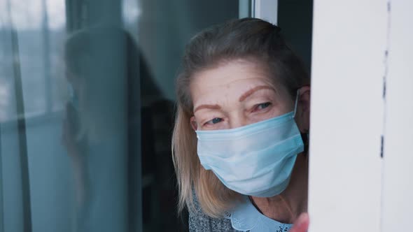 Portrait of Lonely Elderly Woman in Quarantine with Face Mask Looking Through the Window