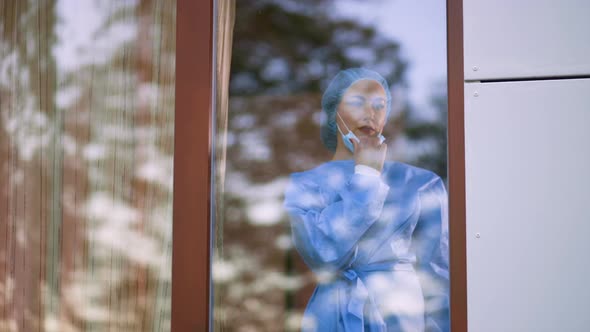 Young Female Doctor or Nurse Takes Off Surgical Face Mask During Home Visit. Behind Window View with