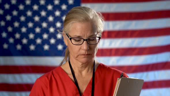 Portrait of nurse set against an out of focus US flag showing disappointment and sadness.