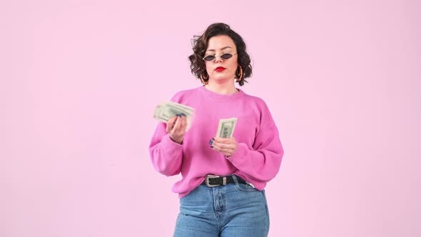 Stylish avarice woman counts a wad dollar money isolated on pink background.