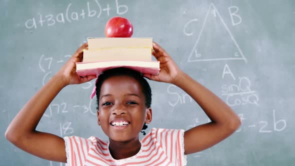 Schoolgirl holding books stack with apple on head against chalkboard