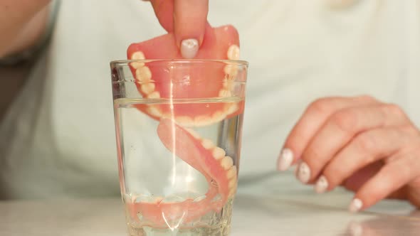 A woman takes from a glass and puts on false teeth while at home.