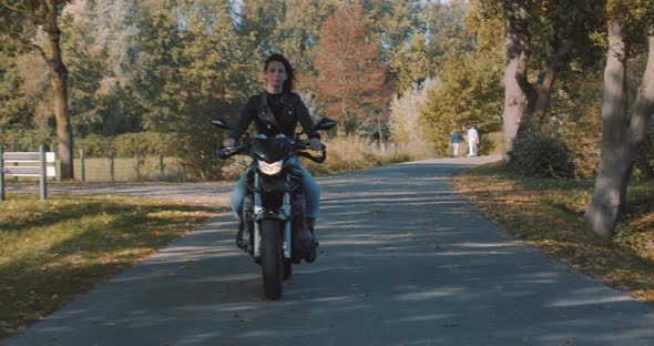 Pretty smiling European young woman driving a motorbike wearing leather jacket in forest with vibran