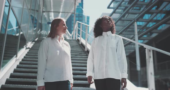 Two young women in business attire look around and talks while standing on the stairs