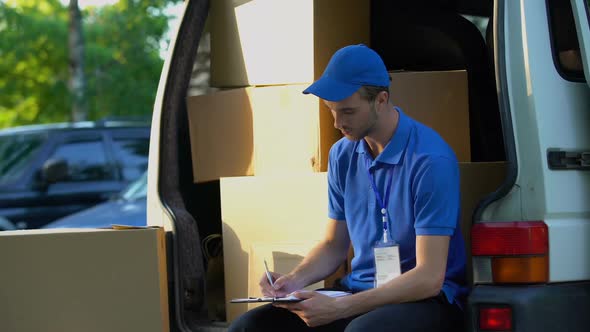 Young Deliveryman Writing Parcel Distribution Report and Smiling, Sitting in Van