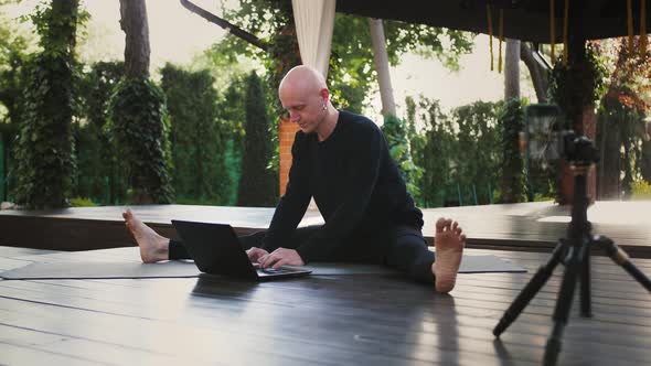 Man in Black Sportswear is Performing Yoga and Typing on Laptop Sitting on Mat in Landscaped Patio