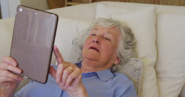 Caucasian senior woman smiling while using digital tablet lying on the couch at home