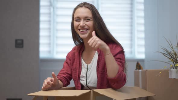 Beautiful Happy Woman Gesturing Thumbs Up Smiling Looking at Camera Sitting in Box Indoors
