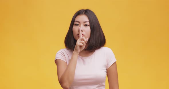 Gesture of Secret. Mysterious Asian Lady Holding Finger on Her Lips, Orange Studio Background.