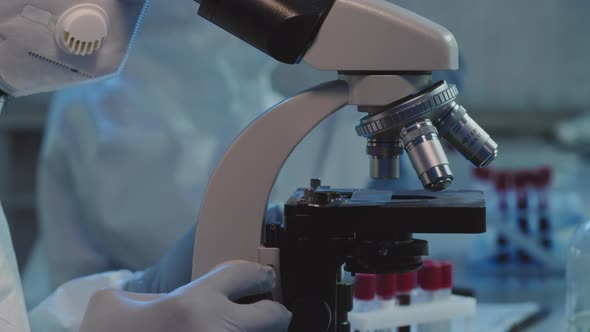 Scientist in Coveralls and Face Mask Studying Sample under Microscope