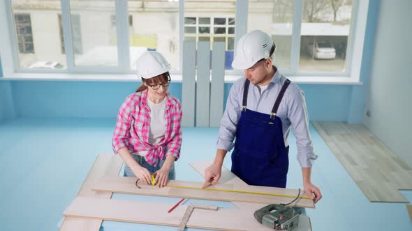 Woman and Man Builtin Hard Hats Measure Laminate with Tape Measure for Renovating Floor in New