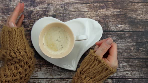 Hands of a Girl in a Knitted Jacket Hugging a Cup of Coffee or Cappuccino in the Shape of a Heart