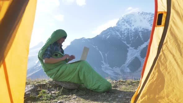 Man in Sleeping Bag Opens Laptop and Types Sitting on Rock