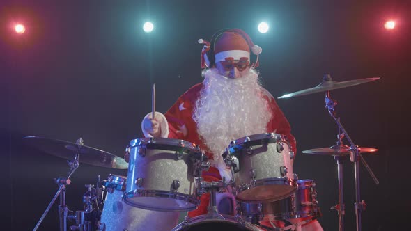 Santa Claus in a Red Costume with Beard and Funny Glasses Plays the Drums in a Dark Studio with