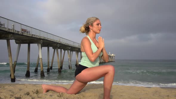 A young attractive woman doing yoga on the beach next to a pier.