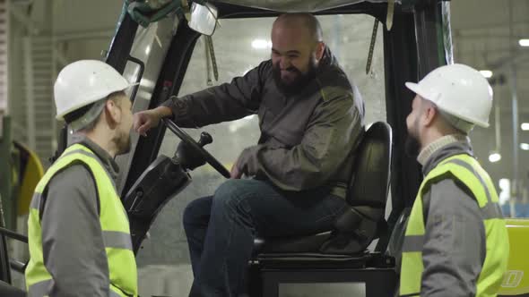 Two Male Caucasian Workers in Helmets and Vests Talking To Bearded Colleague Sitting in Tow Tractor