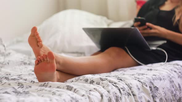 Female sitting on bed with laptop and moving her feet. Woman tapping on her smartphone while moving