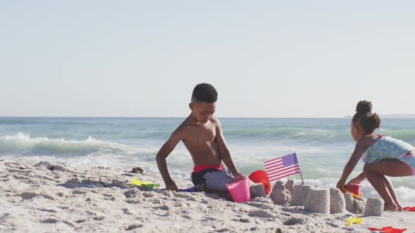 Smiling african american siblings building sandcastle with american flag on sunny beach