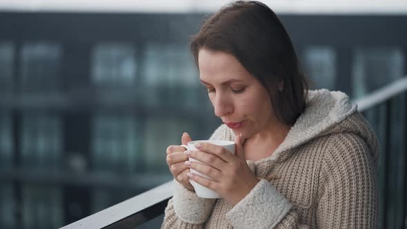 Caucasian Woman Stays on Balcony During Snowfall with Cup of Hot Coffee or Tea