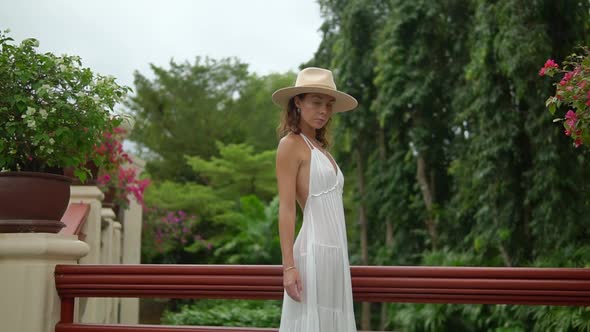 Woman Walks at Resort Area Between Flowers and Trees, She Wears Hat and Dress