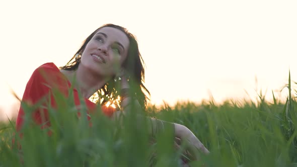 Portrait of a Beautiful Young Hippie Woman in a Red Dress Sits in the Grass of Young Wheat at Sunset