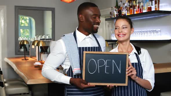 Smiling waiter and waitress holding slate with open sign in restaurant