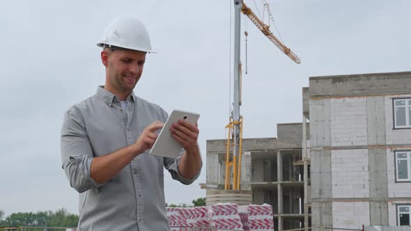 Construction Engineer in a White Helmet with a Yellow Crane in the Background Uses a Tablet