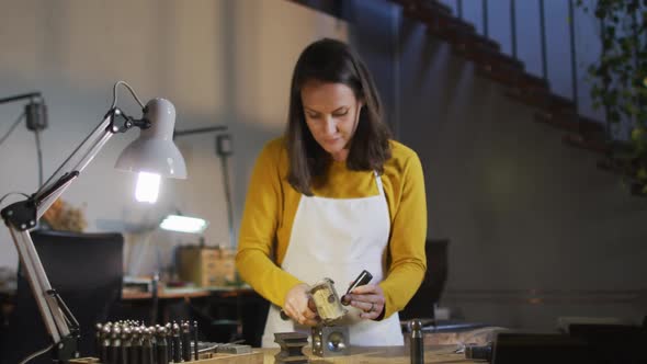 Caucasian female jeweller in workshop wearing apron, using hammer, making jewelry