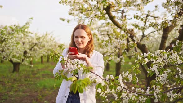 A Woman in a White Jacket Shoots a Video on Her Phone in a Flowering Garden