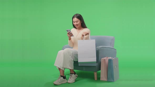 Asian Woman Sitting On Sofa And Holding Shopping Bag Enjoy Using Mobile Phone On Green Screen