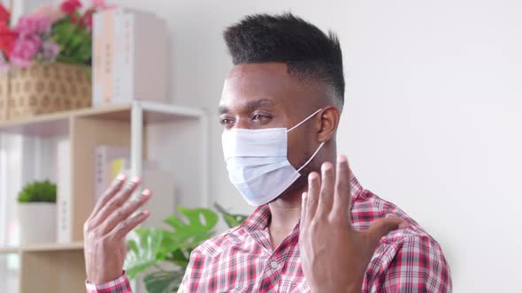 African male wearing mask sitting on a sofa in living room and talking with friend by video call